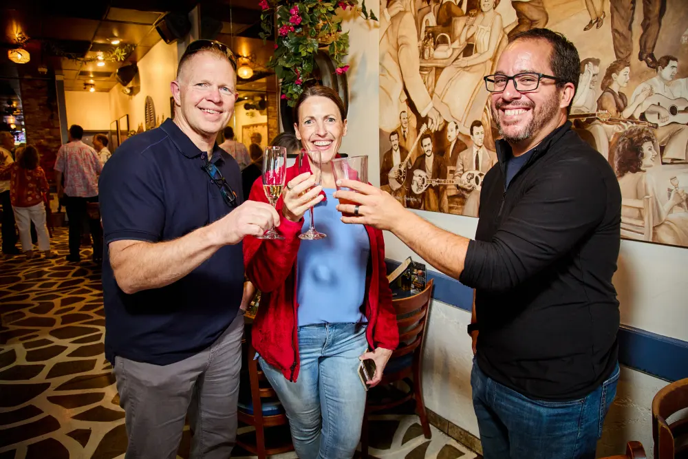 Three guests raising a glass beside the mural at Demitri’s Taverna